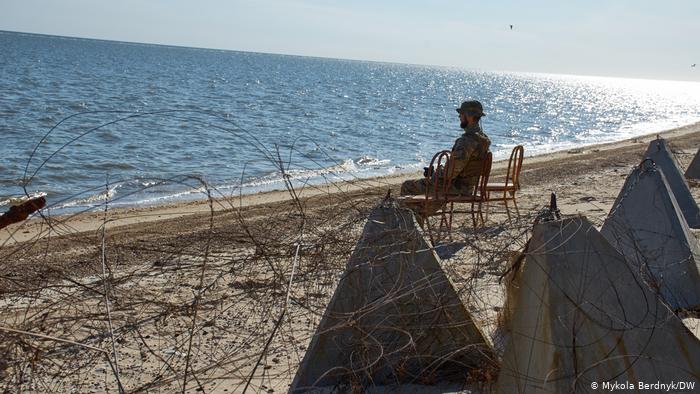 Мужчина сидит на берегу моря - рядом колючая проволока и бетонные заграждения