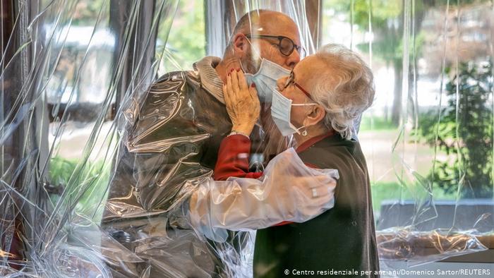 Пожилая женщина, живущая в доме престарелых, обнимается с пришедшим навестить ее сыном через пластиковую штору.