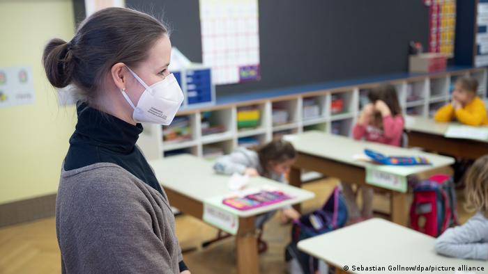 Воспитательница детсада в защитной маске наблюдает за сидящими за столиками детьми